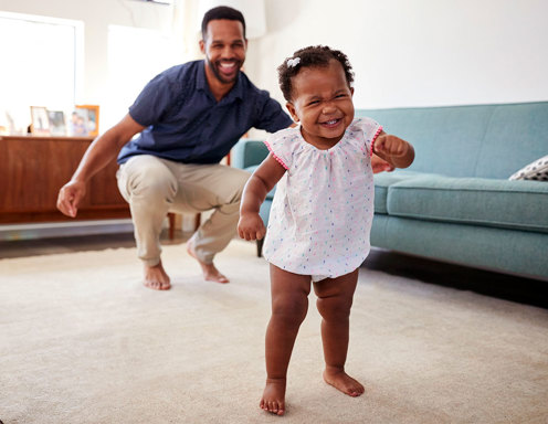 A young toddler girl with dark brown skin and short hair with a bow is smiling as she walks barefoot across the carpet with a young man cheering her on from behind.