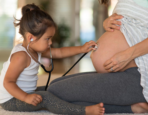 A toddler with light brown skin and hair in a ponytail is holding a stethoscope to a woman's pregnant stomach.