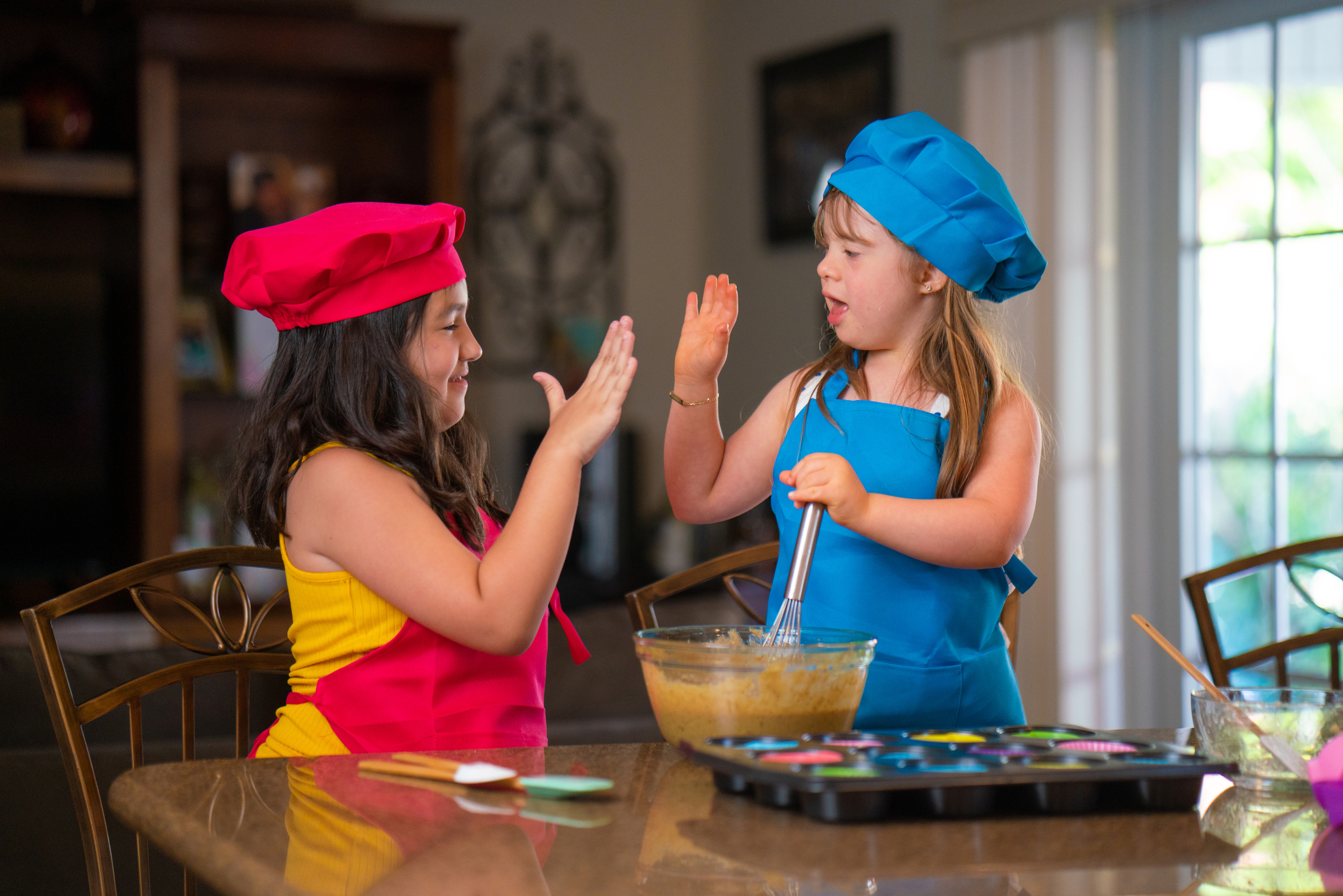 Two girls with light skin and long brown hair both dressed as chefs with a mixing bowl and baking tools on a table in front of them. 