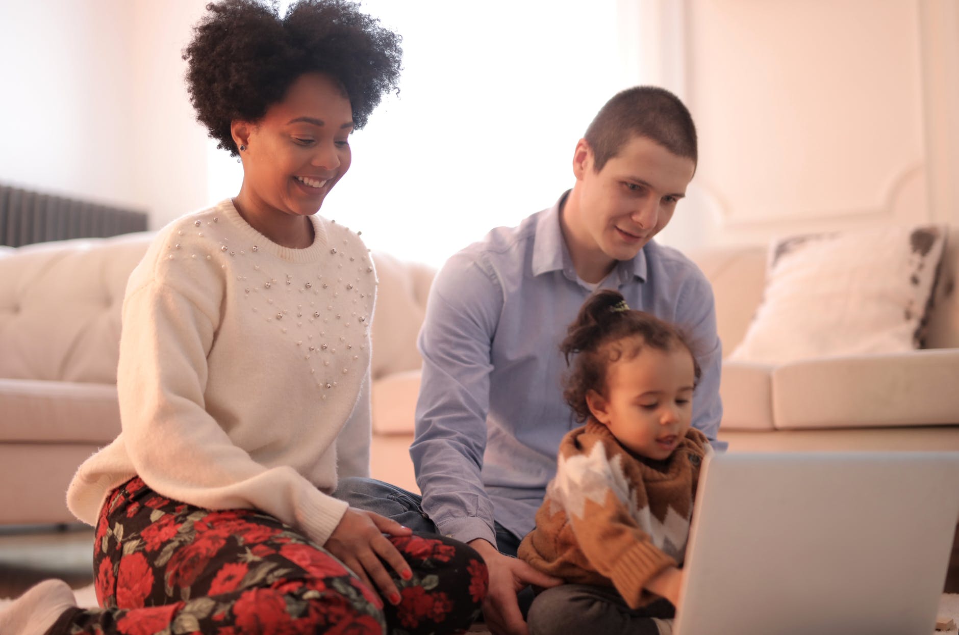 Young women and man sit with toddler on the floor, while all looking at a laptop computer screen. Cozy indoor setting. 