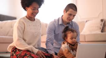 Young women and man sit with toddler on the floor, while all looking at a laptop computer screen. Cozy indoor setting. 