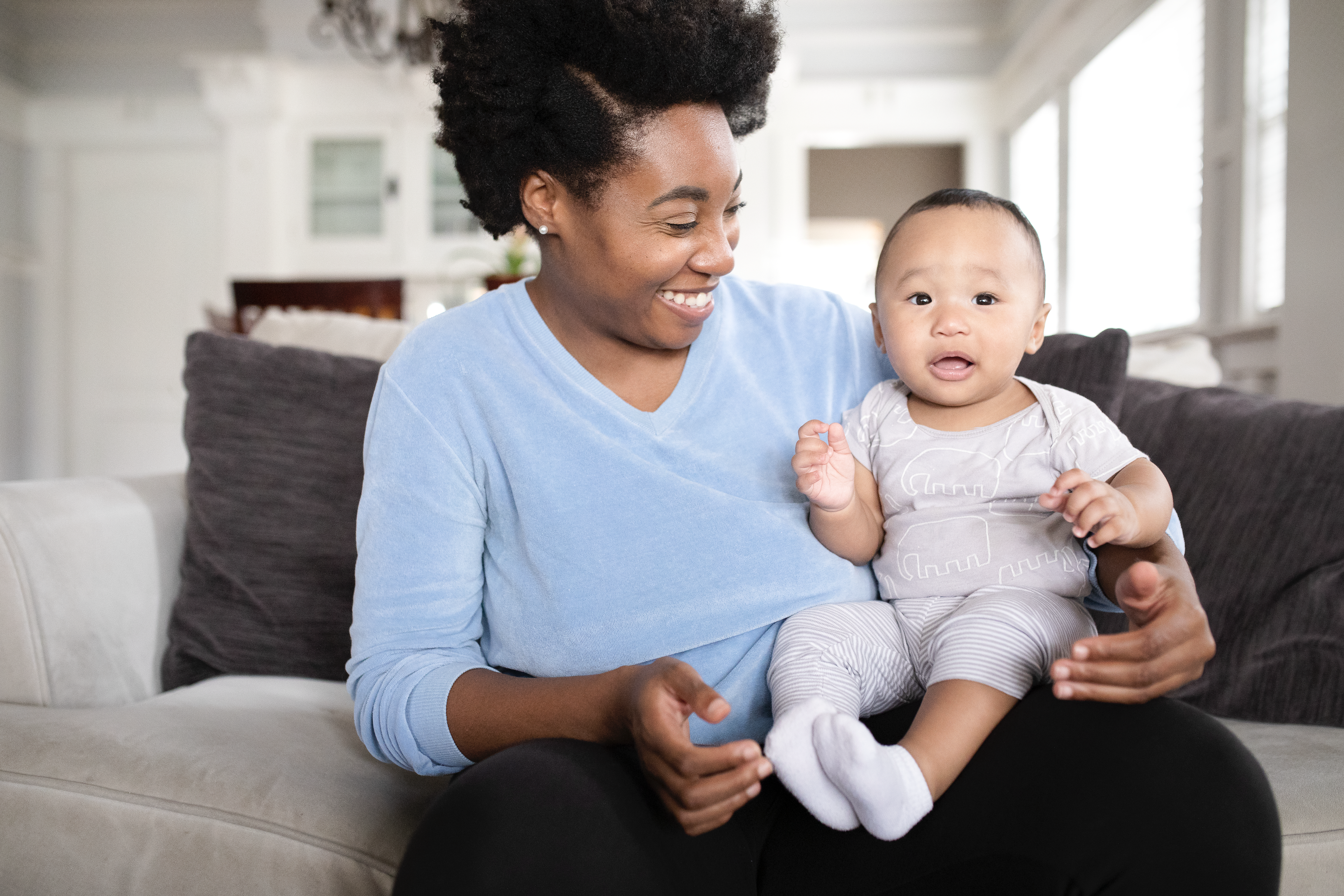 Cheerful African American women smiling down at baby boy in her lap while sitting on the couch. 