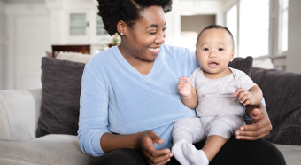 Cheerful African American women smiling down at baby boy in her lap while sitting on the couch. 