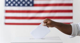 Hand casting a vote into a voting box, with American flag in backdrop 