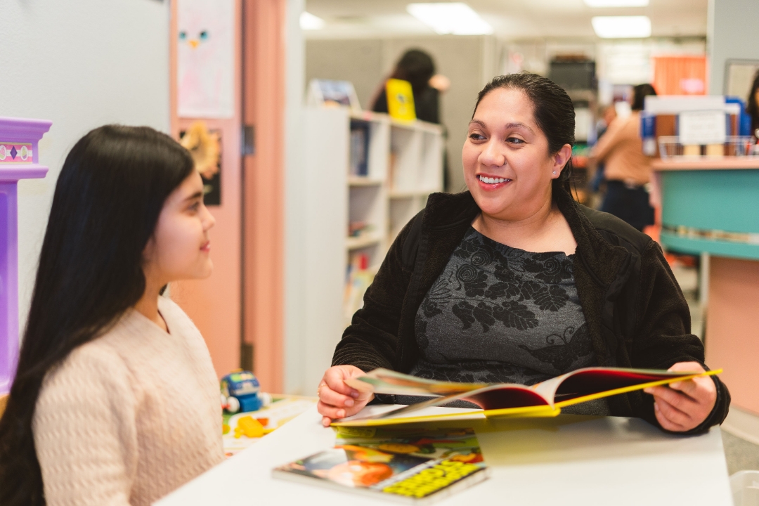 Maria Aguayo Galvan in a classroom setting, smiling and talking to young girl while holding a book