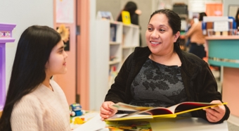 Maria Aguayo Galvan in a classroom setting, smiling and talking to young girl while holding a book