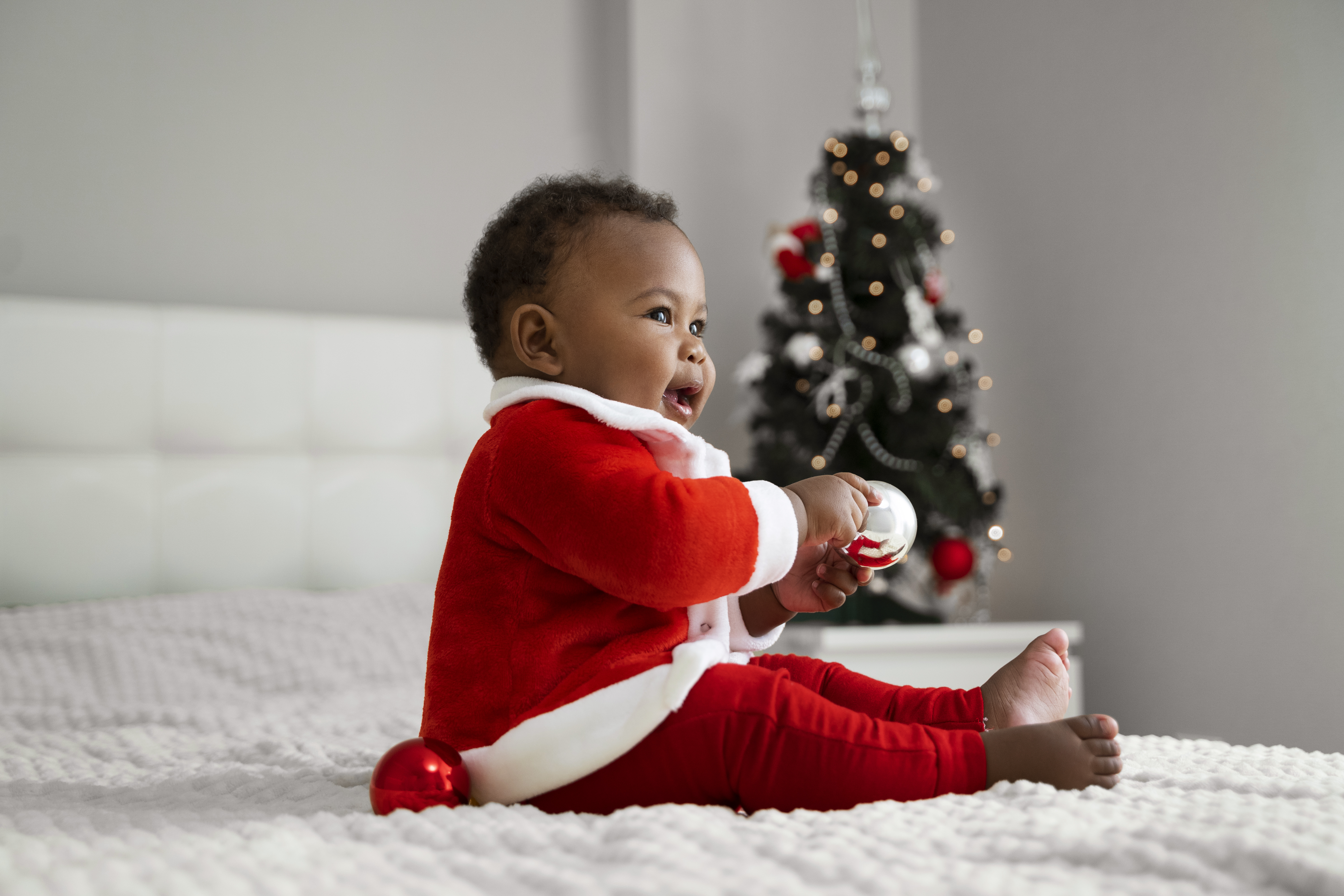 Side View Smiley Baby Indoors with Santa outfit on, white comfy looking bed with Christmas tree in background 