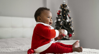 Side View Smiley Baby Indoors with Santa outfit on, white comfy looking bed with Christmas tree in background 