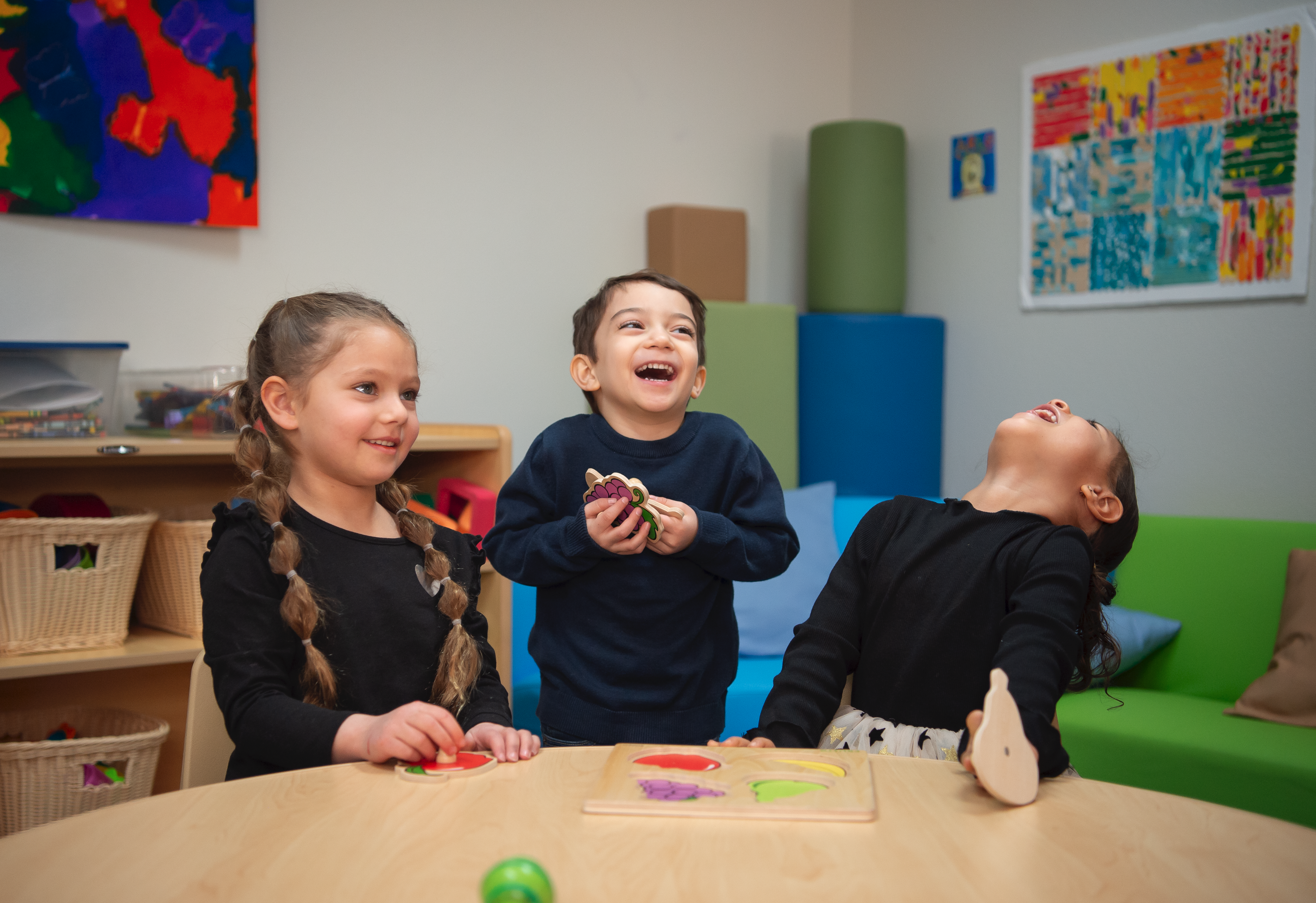 Two little girls and one little boy in a classroom setting laughing while playing with toys