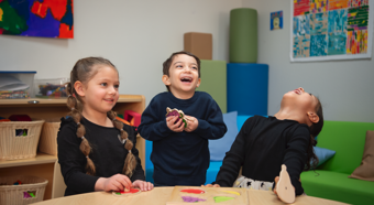 Two little girls and one little boy in a classroom setting laughing while playing with toys