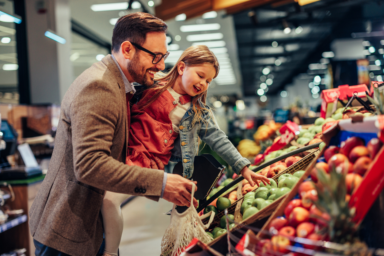 A middle-aged man with light skin and dark hair wearing a business suit is holding a young girl with light skin and long brown hair while they pick out apples at the supermarket together.