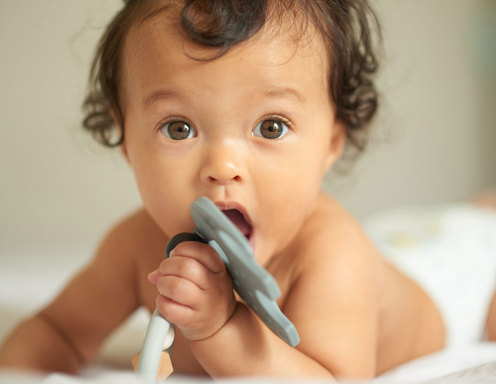 An infant with light skin, curly brown hair and brown eyes is holding a baby toy in its mouth while lying on its stomach in a diaper.