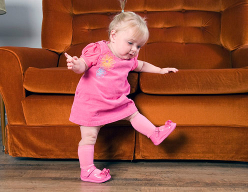 A young toddler-aged girl with light skin and blonde hair in pigtails wearing all pink is holding onto an orange couch while trying to walk.