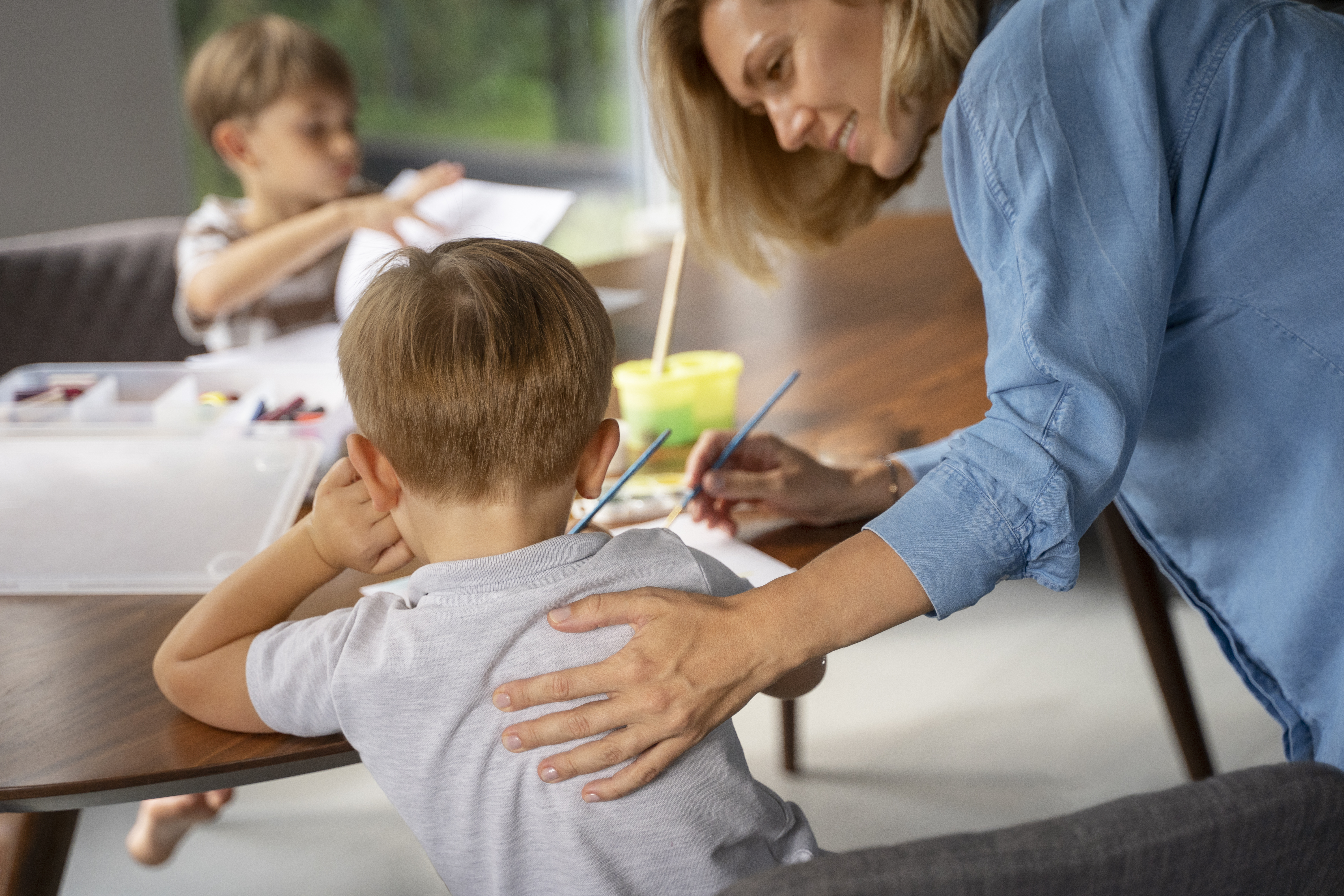 Blonde haired women patting a young boy on the back, working in a classroom setting on an art project