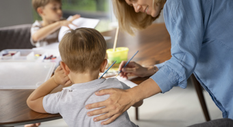 Blonde haired women patting a young boy on the back, working in a classroom setting on an art project