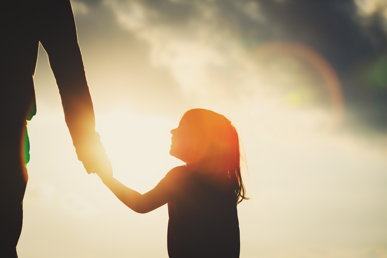 A silhouette-type image of a young girl holding an adult's hand with the sun in the background.