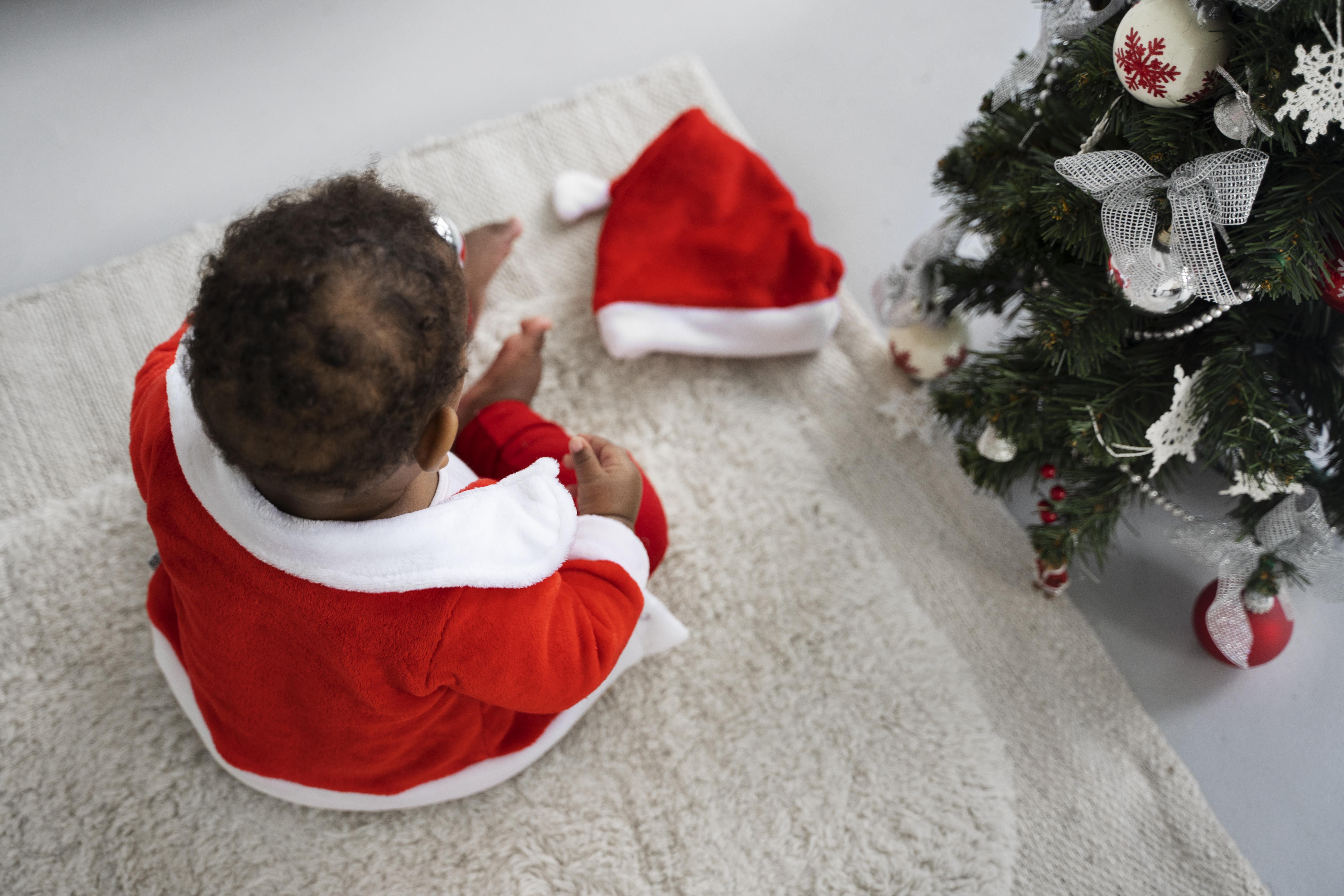 baby playing inside with Santa outfit on next to Christmas tree, high angle