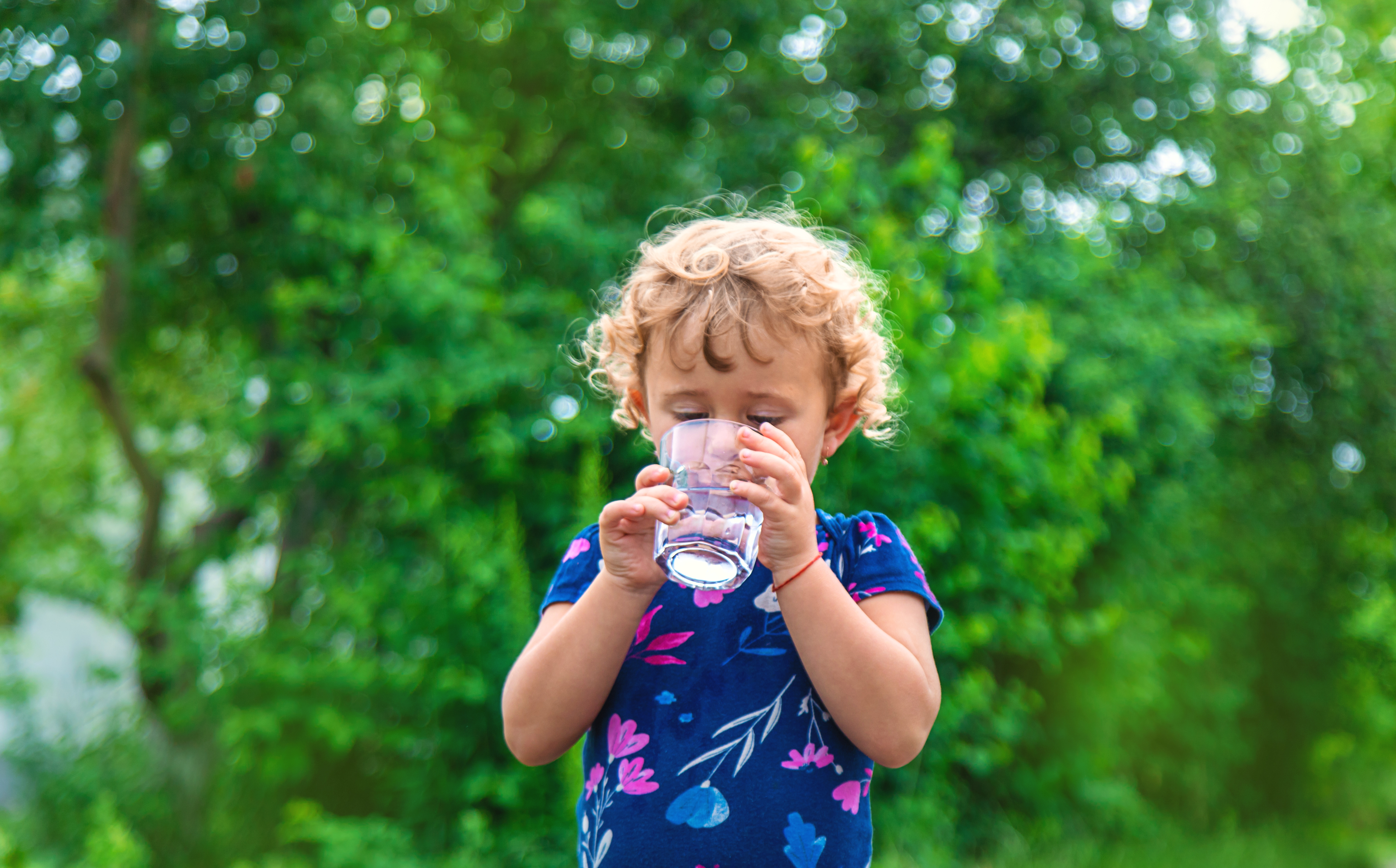 Young child drinks water out of small glass cup while standing outside with a bright green tree backdrop. 