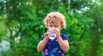 Young child drinks water out of small glass cup while standing outside with a bright green tree backdrop. 