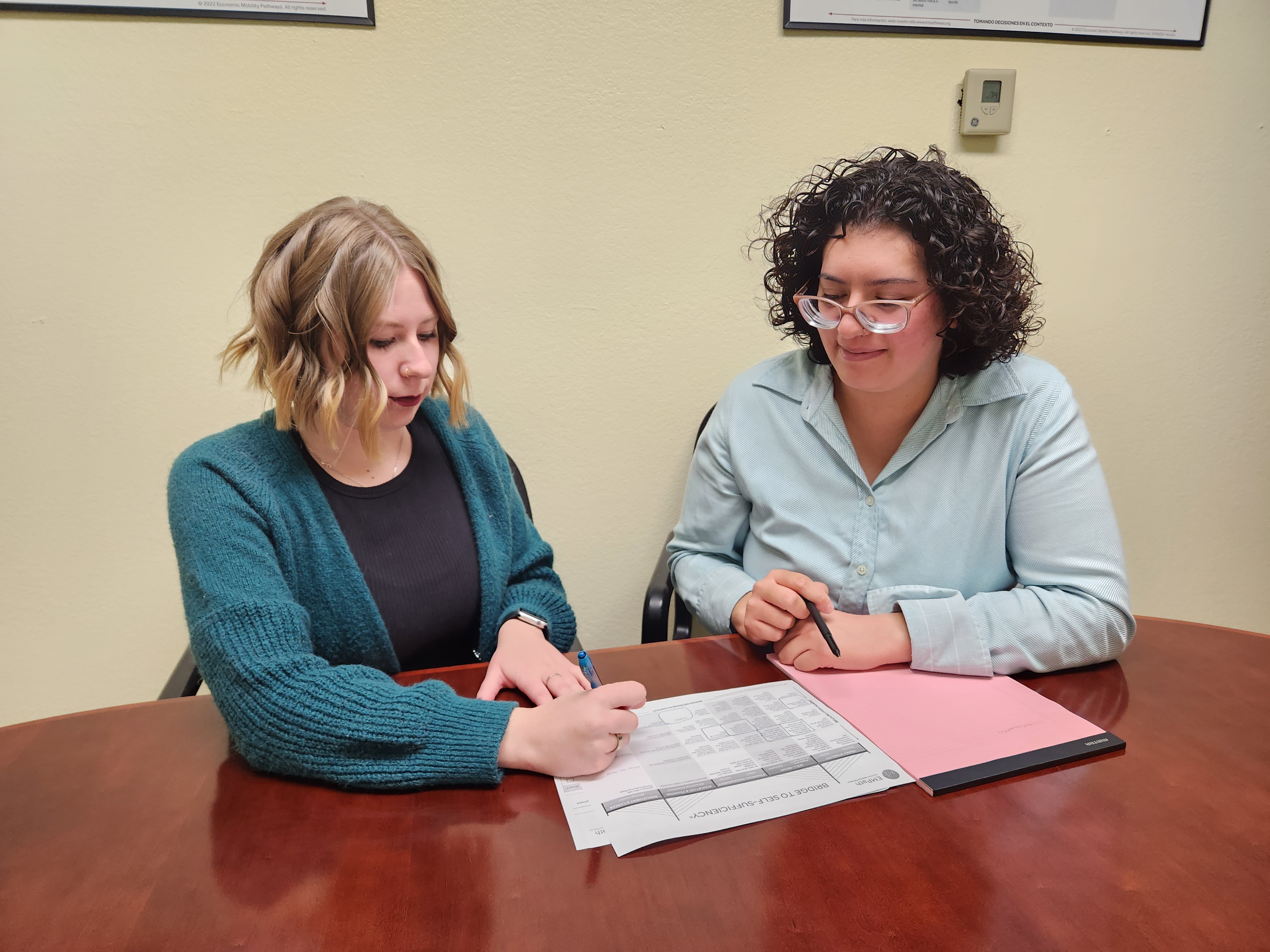 Two middle aged women sitting at a wood desk, while looking over a chart and talking