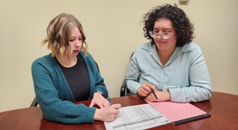 Two middle aged women sitting at a wood desk, while looking over a chart and talking