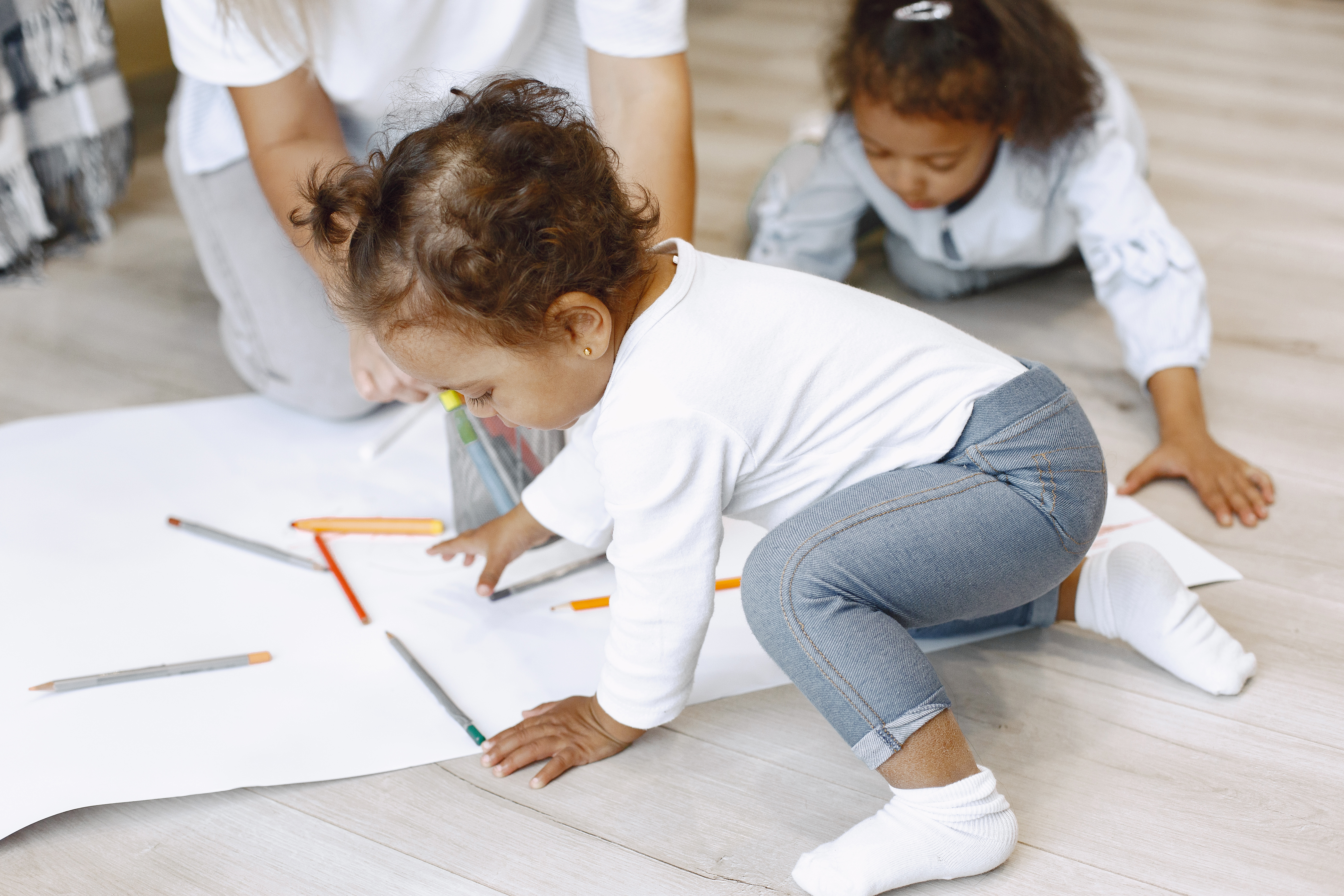 Two toddler girls play on floor, drawing on big pieces of paper, both waring white shirts and blue jeans 