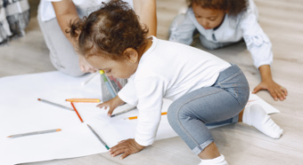 Two toddler girls play on floor, drawing on big pieces of paper, both waring white shirts and blue jeans 